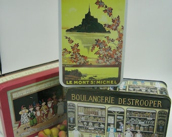 May include: Three vintage tin boxes with colorful illustrations. The top box features a yellow background with a black silhouette of Mont Saint Michel and pink flowers. The middle box has a red background with a black chalkboard and the text "Aa Bb Cc Dd The Christian Pageant". The bottom box has a black background with a colorful illustration of a bakery storefront with the text "Boulangerie Destrooper".
