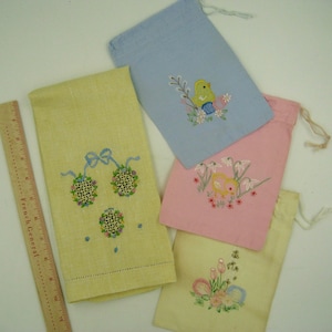 May include: Four embroidered fabric bags with different designs. One bag is light blue with a yellow bird and flowers. Another bag is pink with a yellow chick and white flowers. A third bag is cream with a floral design and two eggs. The fourth bag is yellow with a blue bow and three embroidered circles.