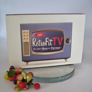 May include: A box featuring a retro-style television design with the text "RetroFit TV" and the tagline "for your iPhone or iPod touch!". The TV is purple with a brown frame and gold legs. A small bunch of colorful artificial fruit is in the foreground.