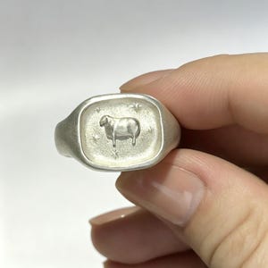 May include: A matte silver signet ring featuring a rectangular face with rounded corners. The center of the ring displays an embossed sheep figure surrounded by four small, etched star shapes on a textured, light-colored metallic surface.