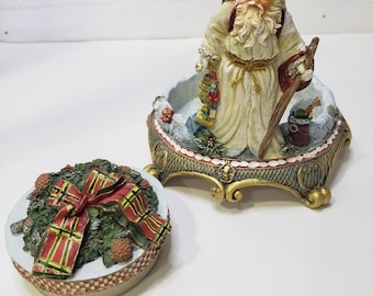May include: A decorative Santa Claus figurine standing inside a circular base with a matching lid. The Santa wears a cream-colored robe and carries a wooden staff. The lid features a green wreath with pinecones and a red and green plaid ribbon.
