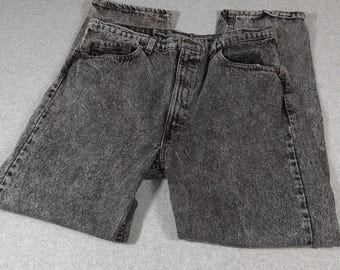 May include: A pair of charcoal gray acid-wash denim jeans featuring a classic five-pocket design, a button fly closure, and a waistband with belt loops. The fabric displays a mottled, textured appearance throughout the legs and pockets.