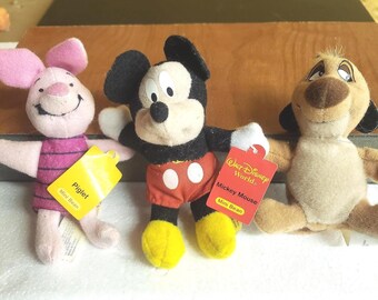 May include: Three plush toys including a pink Piglet with a yellow tag, a Mickey Mouse with a red Walt Disney World tag, and a tan Timon figure. Each character features soft fabric construction and embroidered facial details.