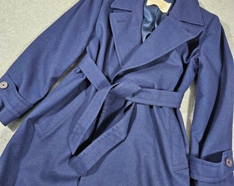 May include: A navy blue long-sleeve trench coat featuring a wide notched collar, a matching fabric tie belt at the waist, and buttoned cuffs. The coat has a smooth texture and includes a visible interior brand label near the neckline.