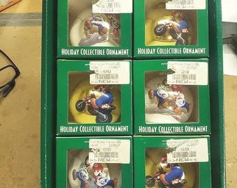 May include: A cardboard box containing six green boxes, each holding a Mario-themed holiday collectible ornament. The ornaments feature Mario characters, some riding motorcycles, and each box has a white price tag with a barcode and the word NEW.