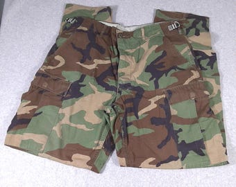 May include: Cargo pants featuring a woodland camouflage pattern in shades of green, brown, and black. The pants include side cargo pockets with flaps, a button fly closure, and adjustable metal side waist buckles for a secure fit.
