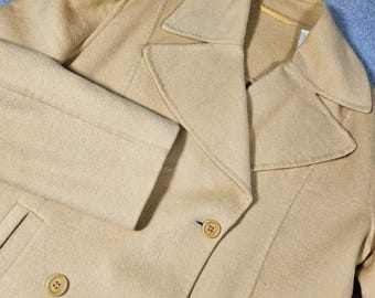 May include: A light tan pea coat featuring a wide notched collar, long sleeves, and a double-breasted front closure with round, matching tan buttons. The garment is constructed from a textured fabric and includes two vertical slit pockets.