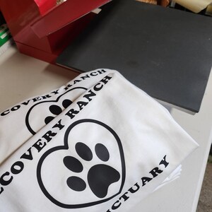Könnte beinhalten: Zwei weiße T-Shirts mit einem schwarzen herzförmigen Pfotenabdruck-Design und den schwarzen Aufdrucken "Discovery Ranch Sanctuary". Ein roter Karton und ein schwarzes quadratisches Objekt befinden sich im Hintergrund.
