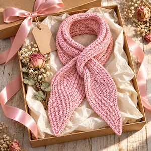 May include: A light pink knitted scarf with a textured stitch pattern, presented in a cardboard gift box lined with white tissue paper, accompanied by a blank brown paper tag and a dried rose with baby's breath flowers.