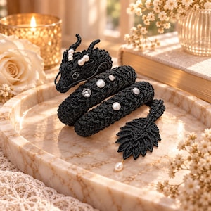 May include: A black crocheted dragon figurine featuring a coiled body with intricate stitch patterns, embellished with small white pearls and clear crystals along its back, and a leaf-shaped tail tip accented with a single dangling pearl.