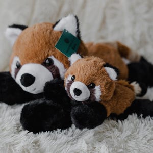 May include: Two plush red panda stuffed animals, one large and one small, resting on a fluffy white surface. The pandas have brown fur, white faces, and black paws. A green tag is attached to the larger panda.