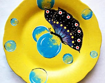 Può includere: Un piatto rotondo giallo con bordo smerlato raffigurante una farfalla nera dipinta a mano con punti bianchi e rossi. Il design include diverse forme circolari blu e gialle simili a bolle sparse sulla superficie.
