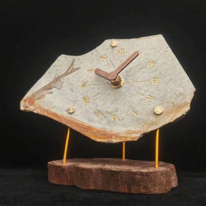 Puede incluir: Un reloj de mesa con una esfera de piedra gris claro irregular que presenta una impresión de pez fosilizado. El reloj tiene números romanos dorados, dos manecillas marrones y se apoya sobre una base de madera natural con tres soportes metálicos dorados.