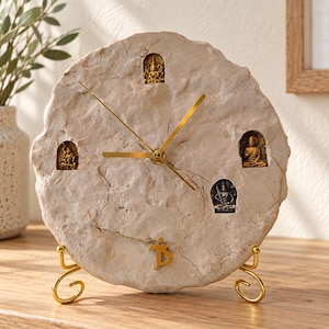 Puede incluir: Un reloj de pared circular hecho de piedra texturizada de color claro con un borde irregular. Presenta tres manecillas delgadas de color dorado y cuatro pequeñas figuras de deidades doradas incrustadas en la esfera. Un símbolo dorado está en la parte inferior.