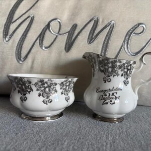 May include: A white ceramic creamer and sugar bowl set with a black floral pattern. The creamer has a silver handle and the text "Congratulations 25th Anniversary" on the front.