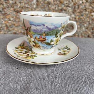 May include: A white teacup and saucer with a gold rim. The teacup features a colourful illustration of a mountain scene with two people in a canoe. The saucer has a similar floral design. The text "Wilford, B.C. British Columbia" is printed on the teacup.