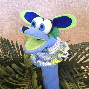 Puede incluir: Una marioneta de calcetín caprichosa con forma de criatura azul y verde. Tiene ojos grandes y expresivos, un pico azul y orejas grandes azules y verdes. La marioneta lleva una bufanda de ganchillo. El fondo incluye follaje verde.
