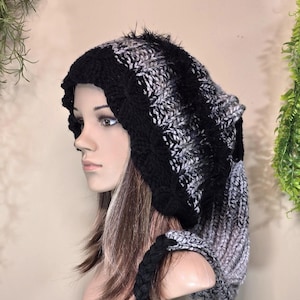 Op de afbeelding: Een zwarte en grijze gebreide capuchon met een lange, vloeiende sjaal eraan vast. De muts is gemaakt van zacht, pluche garen en heeft een slouchy pasvorm.