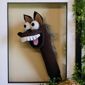 Puede incluir: Un caballo de peluche marrón con ojos grandes y redondos y una amplia sonrisa. El caballo está hecho de fieltro y tiene una larga melena.