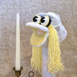 May include: A whimsical sock puppet with large, expressive eyes, a yellow beak, and long, yellow yarn hair. The puppet wears a white lace veil, suggesting a wedding theme. A tall, white candle stands in a brass candlestick to the left.