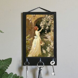 May include: A black key holder with a vintage painting of a woman in a white dress standing in a garden with flowers. The key holder has four hooks for hanging keys.