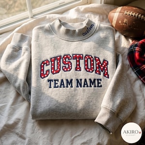 Puede incluir: Una sudadera gris claro de cuello alto con el texto bordado CUSTOM en rojo con lunares blancos y el texto TEAM NAME en azul marino. La palabra custom también está bordada en azul marino en el cuello.