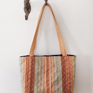 May include: A striped tote bag with brown, orange, and green fabric. The bag has a brown fabric strap and is hanging on a hook.