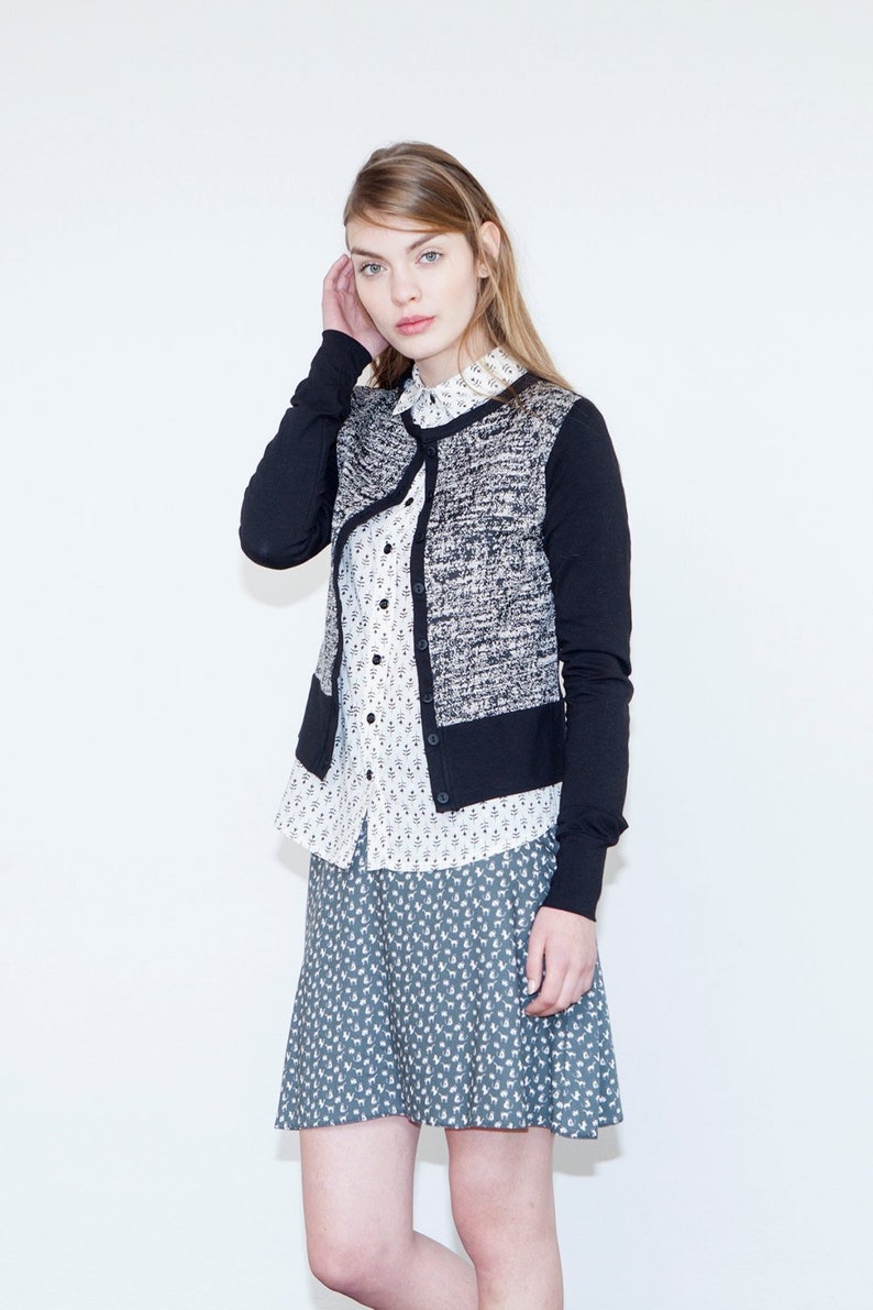 Op de afbeelding: Een vrouw draagt een zwart-witte cardigan over een wit overhemd met knopen en een bloemenprint. Ze draagt ook een rok met een blauw-wit patroon.