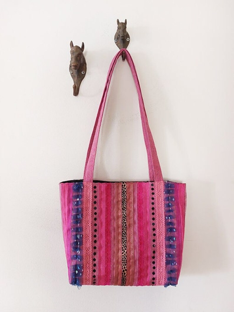 May include: A pink striped tote bag with blue trim and embellishments. The bag has a long strap and is hanging on two hooks.
