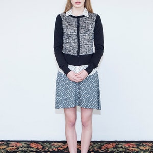 Op de afbeelding: Een vrouw draagt een zwart-witte tweed cardigan over een wit overhemd met kraag, een blauw-witte bloemenrok en beige schoenen. Ze staat op een tapijt met een patroon.