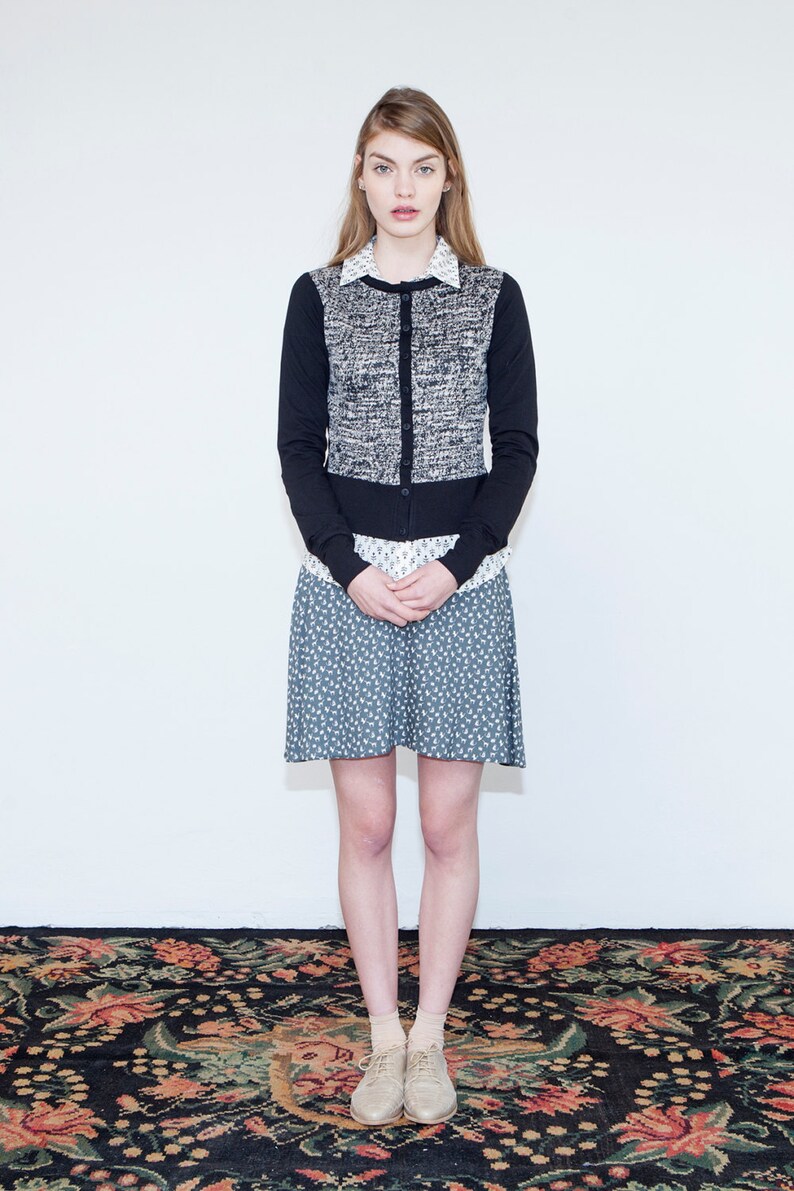 Op de afbeelding: Een vrouw draagt een zwart-witte tweed cardigan over een wit overhemd met kraag, een blauw-witte bloemenrok en beige schoenen. Ze staat op een tapijt met een patroon.
