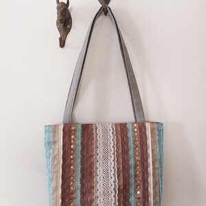 May include: A brown and blue striped tote bag with lace and gold sequins. The bag has two long straps and is hanging on a hook.
