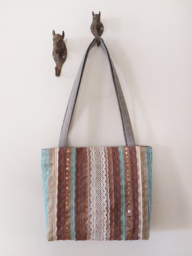May include: A brown and blue striped tote bag with lace and gold sequins. The bag has two long straps and is hanging on a hook.