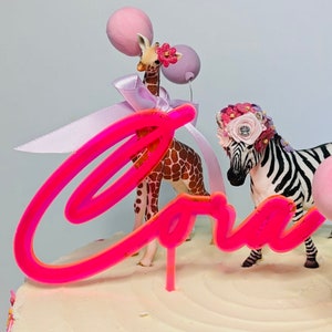 May include: Pink and white cake with a pink neon sign that says "Cora" and a giraffe and zebra figurine on top.