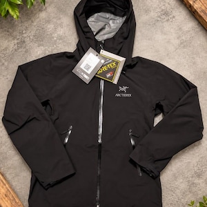 Puede incluir: Una chaqueta negra con capucha Arc'teryx que presenta una cremallera frontal completa, dos bolsillos laterales con cremallera y un forro interior gris. La chaqueta incluye una etiqueta Gore-Tex y el logo de la marca impreso en blanco.