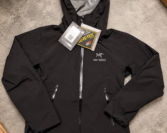 Könnte beinhalten: Eine schwarze Arc'teryx Kapuzenjacke mit durchgehendem Frontreißverschluss, zwei seitlichen Reißverschlusstaschen und grauem Innenfutter. Die Jacke ist mit einem Gore-Tex-Etikett und dem weißen Markenlogo auf der linken Brust versehen.