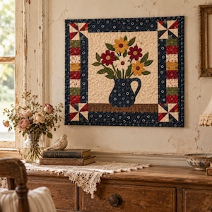 May include: A square fabric wall hanging featuring a central cream-colored panel with an appliqued dark blue pitcher filled with yellow, red, and white flowers. The border consists of dark blue floral fabric and colorful geometric blocks.