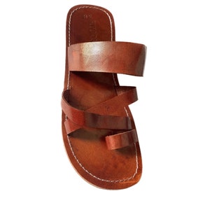 May include: Brown leather sandals with a toe loop and multiple straps. The sandals have a stitched sole and a simple, minimalist design. The leather appears to be smooth and has a natural, warm tone.