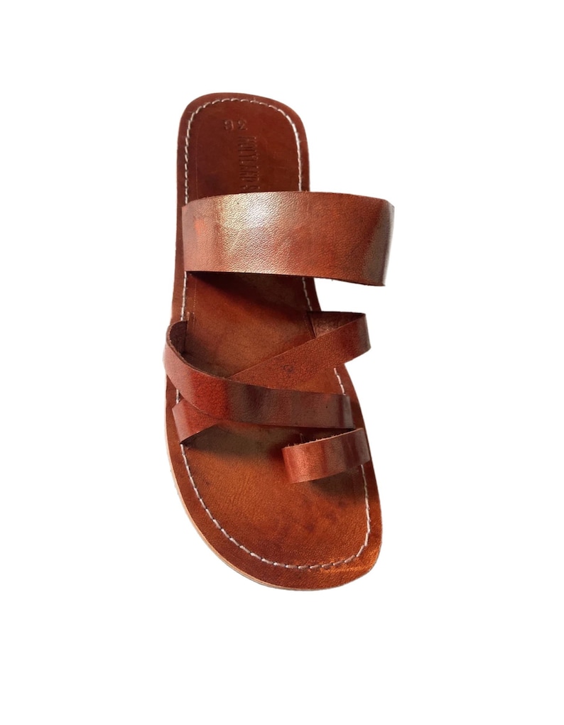 May include: Brown leather sandals with a toe loop and multiple straps. The sandals have a stitched sole and a simple, minimalist design. The leather appears to be smooth and has a natural, warm tone.