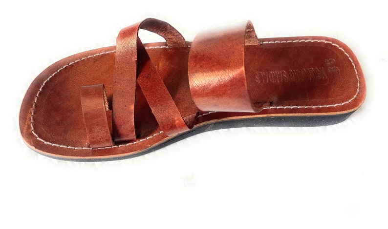 May include: Brown leather sandals with a toe loop and multiple straps. The sandals have a black sole and white stitching around the edges. The brand name "VOLCANO SANDALS" is visible on the footbed.