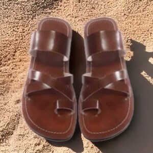 May include: A pair of brown leather sandals with multiple straps and a toe loop. The sandals are set against a sandy background, showcasing their design and construction. The sandals are designed for comfort and style.