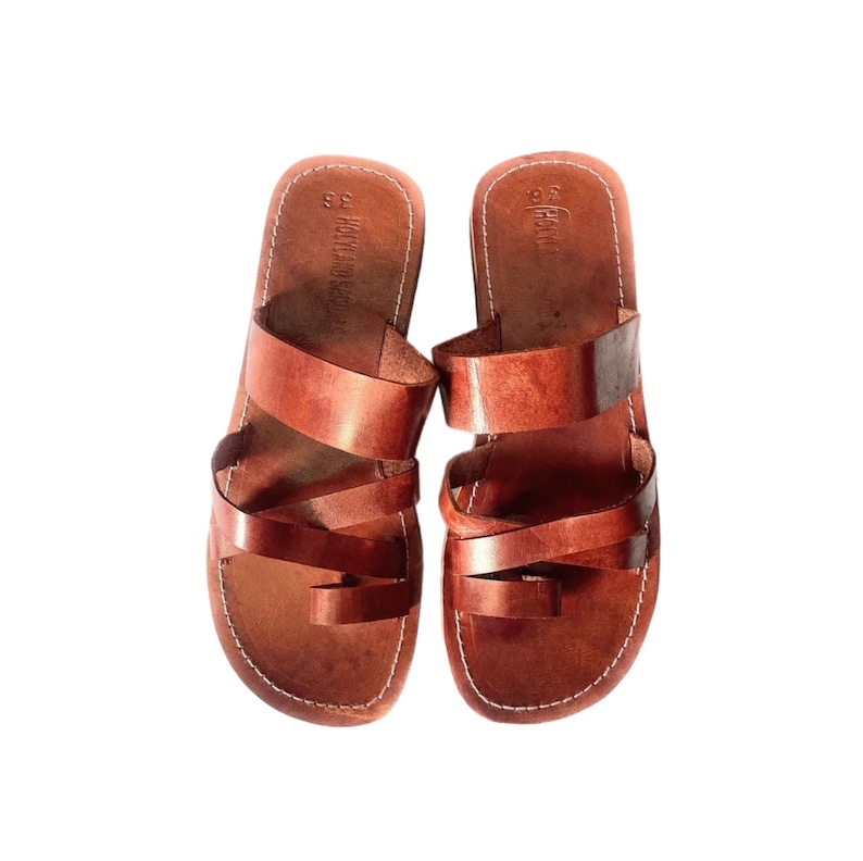 May include: A pair of brown leather sandals with multiple straps and a toe loop. The sandals have a stitched sole and are set against a white background. The sandals are a size 36.