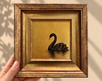 May include: A square framed painting featuring a black swan swimming on a textured gold background. The swan has a red beak and is depicted in profile. The artwork is enclosed in a wide, ornate, metallic gold frame with a distressed finish.