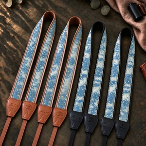 May include: Four camera straps featuring a blue sky pattern with gold stars. Two straps have tan leather ends embossed with the Alex Carter logo, and two straps have black leather ends embossed with the same logo. The straps have scalloped edges.