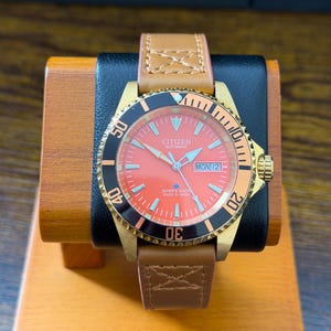May include: A Citizen automatic wristwatch with a bright orange dial, luminous hands, and a day-date window at the three o'clock position. The watch features a gold-tone case, a black rotating bezel with white markers, and a tan leather strap.
