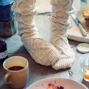 May include: A pair of white knitted socks with a cable knit pattern. The socks are worn by a person sitting on a floor with a cup of coffee, a plate of toast with jam, and a glass of juice.