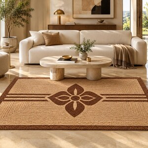 May include: A rectangular woven area rug featuring a natural tan fiber base with a dark brown border and a central four-petaled floral motif. The design includes horizontal dark brown stripes extending from the center to the edges.