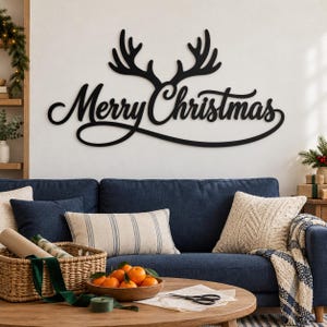 May include: A black wall-mounted sign featuring the text Merry Christmas in a cursive script font, topped with a pair of reindeer antlers. The sign is constructed from a flat material and is mounted on a plain, light-colored wall.