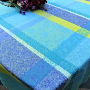 May include: A rectangular tablecloth featuring a woven plaid pattern in shades of light blue, dark blue, and lime green. The fabric displays intricate floral and geometric jacquard motifs within the stripes. A bouquet of artificial flowers rests on top.