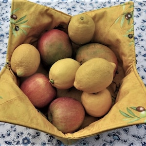 Op de afbeelding: Een gele stoffen kom in stervorm, gevuld met vers fruit: citroenen, appels en mango's. De kom is versierd met olijftakmotieven. Hij staat op een wit tafelkleed met blauwe bloemmotieven.